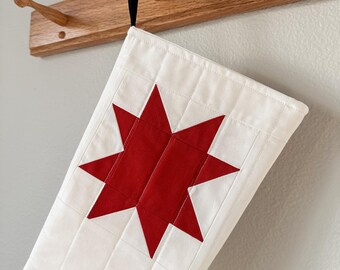 Op de afbeelding: Witte, gestikte kerstsok met een rood stermotief. De sok hangt aan een houten haak.