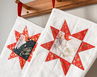 Op de afbeelding: Twee witte kerstsokken met rode en gouden stermotieven. Elke sok heeft een afbeelding van de kerstman en de tekst "Prettige Kerst".