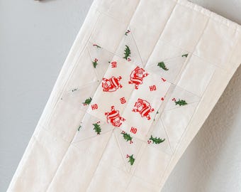 Op de afbeelding: Witte, gewatteerde kerstsok met een stervormige patch met een rode en groene kerstmanprint. De patch zegt meerdere keren "HO".