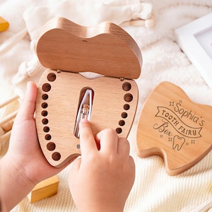 Op de afbeelding: Een houten tandenfee-doosje in de vorm van een tand, met een scharnierend deksel. De doos is open en toont een klein compartiment met een tand. Een apart tandvormig deksel ligt in de buurt, met de tekst "Sophia's Tooth Fairy Box".