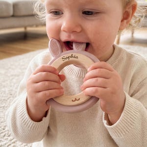 May include: A wooden baby teether shaped like a bunny, with a light purple silicone ring. The teether is engraved with the name "Sophia". The baby is wearing a cream-coloured jumper.