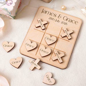 Op de afbeelding: Een lichtgekleurd houten boter-kaas-en-eieren bord gegraveerd met de tekst James & Grace 28th July 2026. Het spel bevat houten X-vormige stukken met het label James en hartvormige stukken met het label Grace, deels op het bord geplaatst.