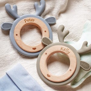 Puede incluir: Dos anillos de dentici&oacute;n circulares con centros de madera clara y bordes de silicona suave con forma de astas de ciervo. Un anillo es azul claro con el nombre Peter grabado y el otro es verde salvia con el nombre Kelly grabado.