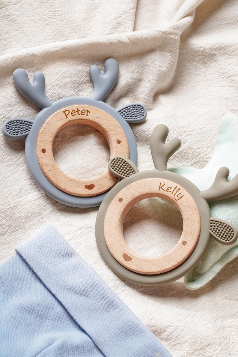 Puede incluir: Dos anillos de dentici&oacute;n circulares con centros de madera clara y bordes de silicona suave con forma de astas de ciervo. Un anillo es azul claro con el nombre Peter grabado y el otro es verde salvia con el nombre Kelly grabado.