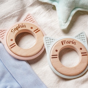 Puede incluir: Dos anillos de dentici&oacute;n circulares con centros de madera clara y bordes de silicona en forma de orejas de gato en rosa suave y azul claro. Los centros de madera est&aacute;n grabados con los nombres Sophia y Maria y detalles de cara de gato.