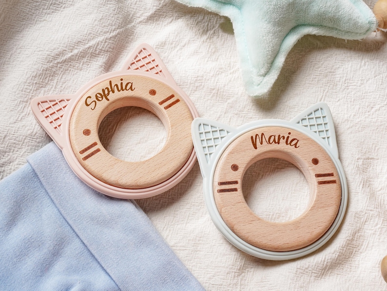 Puede incluir: Dos anillos de dentici&oacute;n circulares con centros de madera clara y bordes de silicona en forma de orejas de gato en rosa suave y azul claro. Los centros de madera est&aacute;n grabados con los nombres Sophia y Maria y detalles de cara de gato.