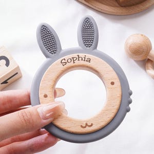 Puede incluir: Un mordedor circular para beb&eacute;s con un marco de silicona gris en forma de orejas de conejo con superficie texturizada y un inserto de madera lisa. El anillo de madera est&aacute; grabado con el nombre Sophia y un peque&ntilde;o dibujo lineal de una boca.