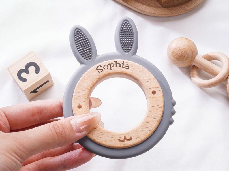 Puede incluir: Un mordedor circular para beb&eacute;s con un marco de silicona gris en forma de orejas de conejo con superficie texturizada y un inserto de madera lisa. El anillo de madera est&aacute; grabado con el nombre Sophia y un peque&ntilde;o dibujo lineal de una boca.