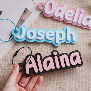 May include: Three custom name keychains featuring raised lettering. One is pink with white text reading Odelia, one is light blue with white text reading Joseph, and one is black with light pink text reading Alaina. Each includes a metal wire loop.