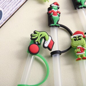 May include: A collection of clear plastic straws with green and red Christmas-themed character decorations. The decorations include a green hand holding a red ornament, and several Grinch figures wearing Santa hats and red coats.