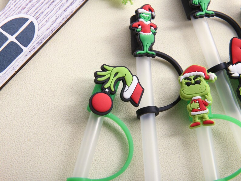 May include: A collection of clear plastic straws with green and red Christmas-themed character decorations. The decorations include a green hand holding a red ornament, and several Grinch figures wearing Santa hats and red coats.