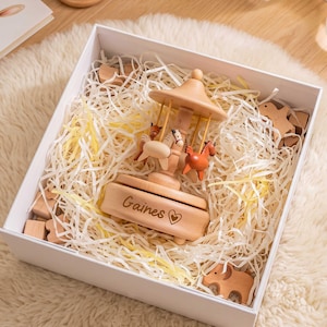 May include: A light-colored wooden carousel music box featuring small horse figures and the engraved name Gaines with a heart symbol. The item is nestled inside a white gift box filled with shredded paper filler and scattered wooden animal shapes.