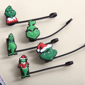 May include: A collection of green Grinch-themed charms with black straps and loops. Some charms feature the Grinch in a Santa hat and red suit. The charms are made of a rubbery material and are likely for use as zipper pulls or similar accessories.