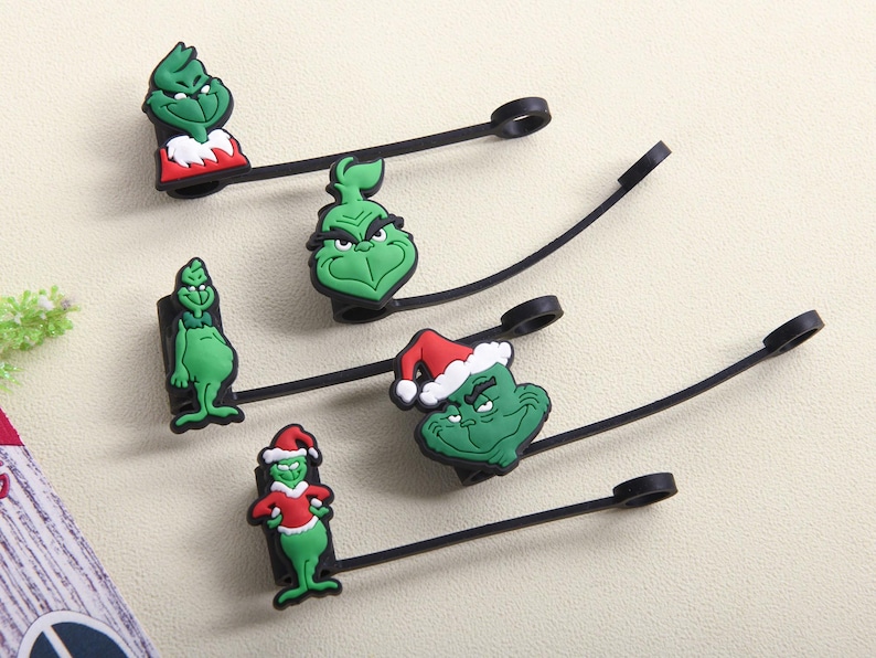 May include: A collection of green Grinch-themed charms with black straps and loops. Some charms feature the Grinch in a Santa hat and red suit. The charms are made of a rubbery material and are likely for use as zipper pulls or similar accessories.