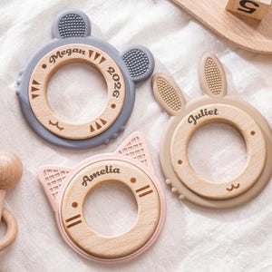 Puede incluir: Tres mordedores circulares para beb&eacute;s con centros de madera y bordes de silicona con forma de orejas de animal. El mordedor gris est&aacute; grabado con Megan 2026, el beige con Juliet y el rosa con Amelia. Tambi&eacute;n se ve un sonajero de madera.