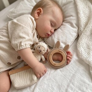 Puede incluir: Un juguete de cordero tejido en crochet beige y un mordedor circular de silicona beige con detalles de madera y orejas de conejo. El mordedor tiene grabado el nombre Jeffery. Se sostiene una peque&ntilde;a bolsa de tela beige con borde de madera.