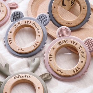 Puede incluir: Una colecci&oacute;n de anillos de dentici&oacute;n circulares con un centro de madera rodeado por un borde de silicona con orejas de animal. Los anillos de madera est&aacute;n grabados con nombres como Chloe, Lauren, Rachel y Olivia, junto con el a&ntilde;o 2026.