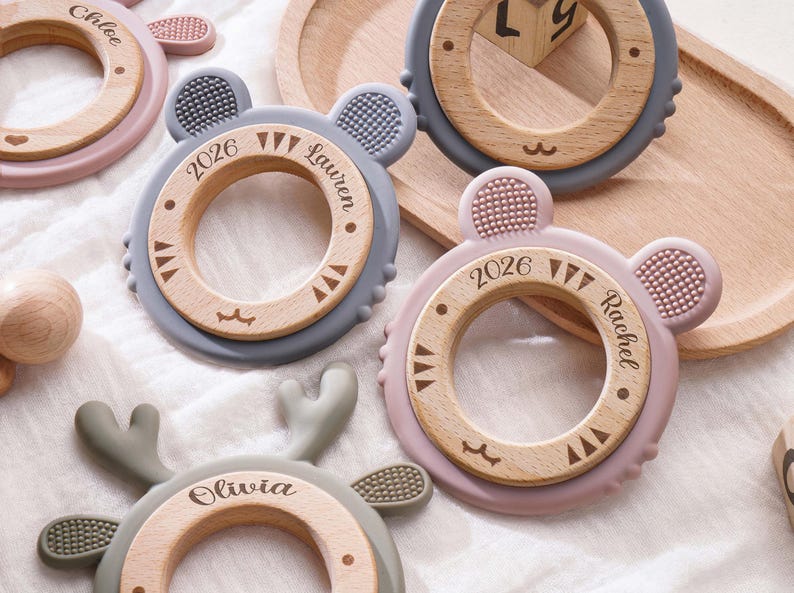 Puede incluir: Una colecci&oacute;n de anillos de dentici&oacute;n circulares con un centro de madera rodeado por un borde de silicona con orejas de animal. Los anillos de madera est&aacute;n grabados con nombres como Chloe, Lauren, Rachel y Olivia, junto con el a&ntilde;o 2026.