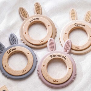 Puede incluir: Cuatro anillos de dentici&oacute;n circulares con centros de madera y orejas de conejo de silicona en gris, beige, crema y rosa empolvado. Cada anillo est&aacute; personalizado con los nombres Ashley, Lauren, Olivia y Hannah grabados en la madera.