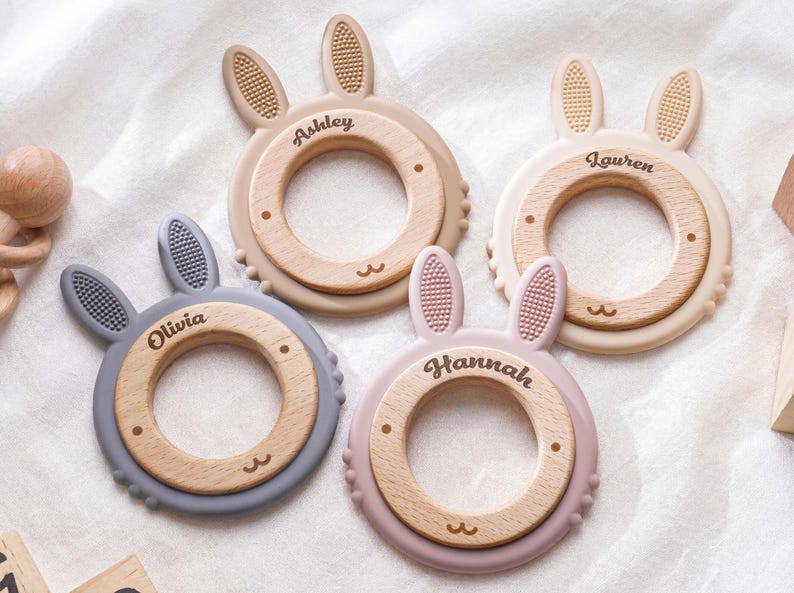 Puede incluir: Cuatro anillos de dentici&oacute;n circulares con centros de madera y orejas de conejo de silicona en gris, beige, crema y rosa empolvado. Cada anillo est&aacute; personalizado con los nombres Ashley, Lauren, Olivia y Hannah grabados en la madera.