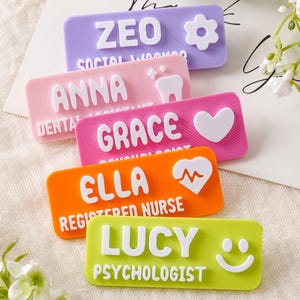 Op de afbeelding: Vijf rechthoekige naamplaatjes in paars, roze, magenta, oranje en limoengroen. Elk plaatje heeft witte verhoogde letters met een naam en functietitel, waaronder ZEO, ANNA, GRACE, ELLA en LUCY, vergezeld van iconen zoals een bloem, tand, hart, hartslag en smiley.