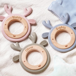 Puede incluir: Tres anillos de dentici&oacute;n circulares con un centro de madera y un marco exterior de silicona con forma de ciervo con astas y orejas. Cada anillo est&aacute; personalizado con un nombre: Nancy en rosa, Sophia en azul y Oliver en verde salvia.