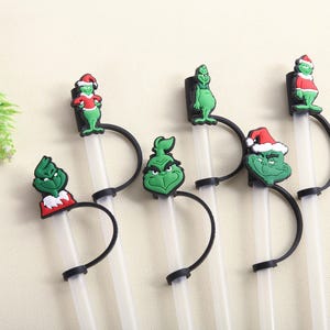 May include: A set of clear plastic straws with black holders and green Grinch-themed toppers. The toppers feature the Grinch in various poses, some wearing Santa hats. Festive and fun for holiday drinks.