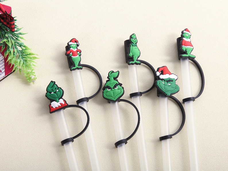 May include: A set of clear plastic straws with black holders and green Grinch-themed toppers. The toppers feature the Grinch in various poses, some wearing Santa hats. Festive and fun for holiday drinks.
