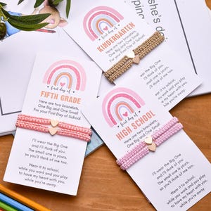 May include: Three sets of two woven bracelets on white cards with rainbow graphics and text reading First Day of Kindergarten, Fifth Grade, and High School. Each set includes two small gold heart charms on adjustable cord bands in tan and pink.