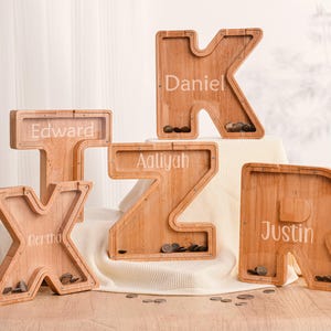 Puede incluir: Huchas de madera con forma de letras, grabadas con nombres. Las letras son K, T, X, Z y R, con los nombres Daniel, Edward, Bertha, Aaliyah y Justin. Las huchas contienen monedas.