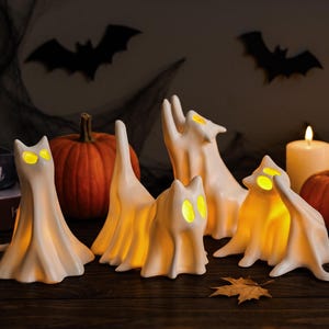 May include: A set of four white, ghost-shaped cat figurines with glowing yellow eyes. Each figurine features a smooth, draped fabric-like appearance, with internal lighting that illuminates the eyes and the body from within, creating a soft, warm glow.