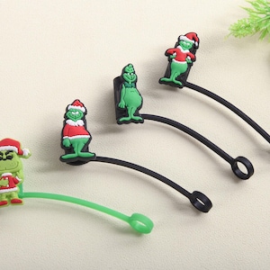 May include: A collection of Christmas-themed charms featuring a green character with a red and white Santa hat and outfit. The charms are attached to black and green rubber loops.