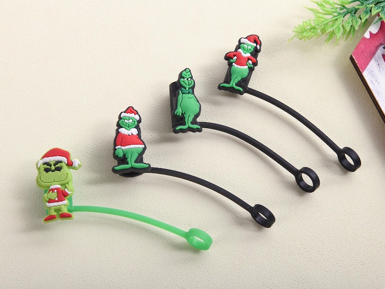 May include: A collection of Christmas-themed charms featuring a green character with a red and white Santa hat and outfit. The charms are attached to black and green rubber loops.
