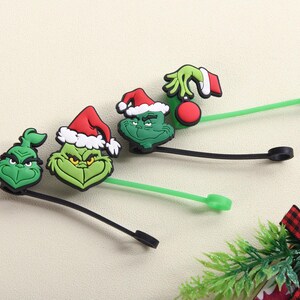 May include: Green Grinch-themed silicone cable ties. Each tie features a different Grinch design, including his face and hand holding an ornament. The ties have black and green stems and are perfect for organizing cables.