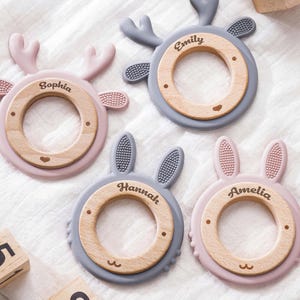 Puede incluir: Cuatro anillos de dentici&oacute;n circulares con centros de madera y bordes de silicona. Dos anillos tienen astas de ciervo y dos tienen orejas de conejo. Los centros de madera est&aacute;n grabados con los nombres Sophia, Emily, Hannah y Amelia. La silicona es rosa y gris.
