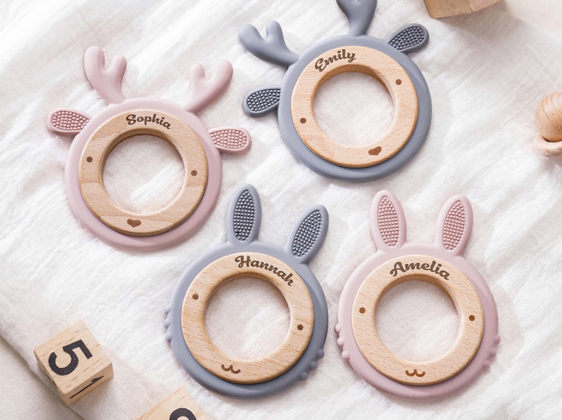 Puede incluir: Cuatro anillos de dentici&oacute;n circulares con centros de madera y bordes de silicona. Dos anillos tienen astas de ciervo y dos tienen orejas de conejo. Los centros de madera est&aacute;n grabados con los nombres Sophia, Emily, Hannah y Amelia. La silicona es rosa y gris.