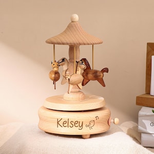Pode incluir: Uma caixa de música de carrossel de madeira com três figuras de cavalos. O carrossel tem um acabamento em madeira clara e apresenta o nome "Kelsey" gravado na base. Um pequeno pássaro e notas musicais também estão gravados.