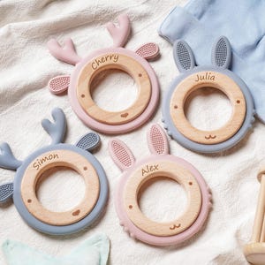 Puede incluir: Cuatro mordedores circulares para beb&eacute;s con centros de madera y bordes de silicona con forma de astas de ciervo u orejas de conejo. Cada mordedor est&aacute; grabado con un nombre: Cherry, Julia, Simon o Alex. Los colores son rosa y azul apagado.