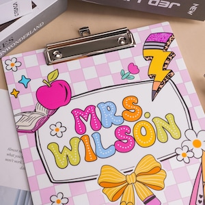 Könnte beinhalten: Ein Klemmbrett mit hellrosa-weißem Karomuster und dem Text "MRS. WILSON" in bunten Buchstaben. Das Design zeigt Illustrationen eines rosa Apfels, eines offenen Buches, eines gelben Blitzes und einer gelben Schleife.
