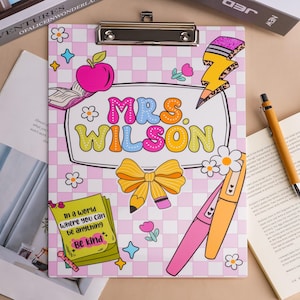 Könnte beinhalten: Ein Klemmbrett mit hellrosa-weißem Karomuster und dem Text "MRS. WILSON" in bunten Buchstaben. Das Design enthält Illustrationen eines rosa Apfels, einer gelben Schleife, eines Blitzes und einer grünen Notiz mit der Aufschrift "Be Kind".