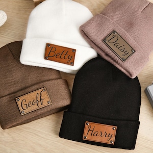Peut inclure: Cinq bonnets en tricot de couleurs blanche, marron, noire et mauve, chacun avec un patch en similicuir affichant un nom : "Betty", "Geoff", "Harry" et "Daisy". Un jouet en bois et un lapin en peluche sont également visibles.