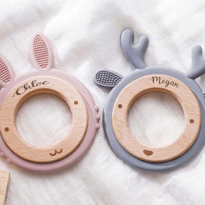 Puede incluir: Dos anillos de dentici&oacute;n circulares con centros de madera y bordes de silicona. El anillo rosa tiene orejas de conejo y el nombre Chloe grabado. El anillo gris tiene astas de ciervo y el nombre Megan grabado. Detalles de silicona texturizada.