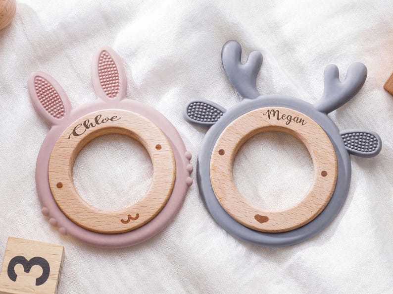 Puede incluir: Dos anillos de dentici&oacute;n circulares con centros de madera y bordes de silicona. El anillo rosa tiene orejas de conejo y el nombre Chloe grabado. El anillo gris tiene astas de ciervo y el nombre Megan grabado. Detalles de silicona texturizada.