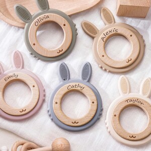 Puede incluir: Cinco anillos de dentici&oacute;n circulares con centros de madera y orejas de conejo de silicona en tonos gris, beige, rosa empolvado, azul y crema. Cada centro de madera est&aacute; grabado con un nombre: Olivia, Alexis, Emily, Cathy y Taylor.