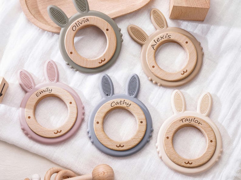 Puede incluir: Cinco anillos de dentici&oacute;n circulares con centros de madera y orejas de conejo de silicona en tonos gris, beige, rosa empolvado, azul y crema. Cada centro de madera est&aacute; grabado con un nombre: Olivia, Alexis, Emily, Cathy y Taylor.
