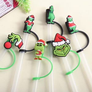 May include: A collection of clear plastic straws with green and black accents, each adorned with a different Grinch-themed character. The Grinch figures wear Santa hats and red and white outfits, perfect for Christmas.