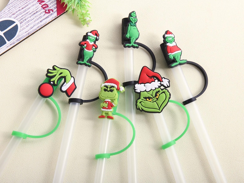 May include: A collection of clear plastic straws with green and black accents, each adorned with a different Grinch-themed character. The Grinch figures wear Santa hats and red and white outfits, perfect for Christmas.