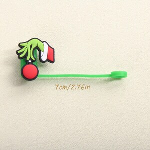 May include: A green, red, and white Christmas-themed charm. The charm features a green hand holding a red ornament. The charm is attached to a green strap. The text "7cm/2.76in" is printed on the image.