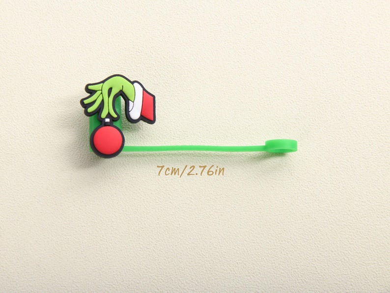 May include: A green, red, and white Christmas-themed charm. The charm features a green hand holding a red ornament. The charm is attached to a green strap. The text "7cm/2.76in" is printed on the image.
