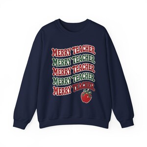 May include: A navy blue long-sleeve sweatshirt featuring the text MERRY TEACHER repeated five times in alternating red and green wavy lines. A red apple illustration with a green bow sits below the final line of text.